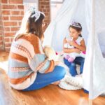 Beautiful teacher and toddler wearing princess crown playing with unicorn doll inside tipi around lots of toys at kindergarten