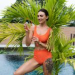 Beautiful european women in two pieces orange bikini swimsuit in perfect shape, good body, tanned, with trendy sunglasses outside at tropical swimming pool and palm leaf tree