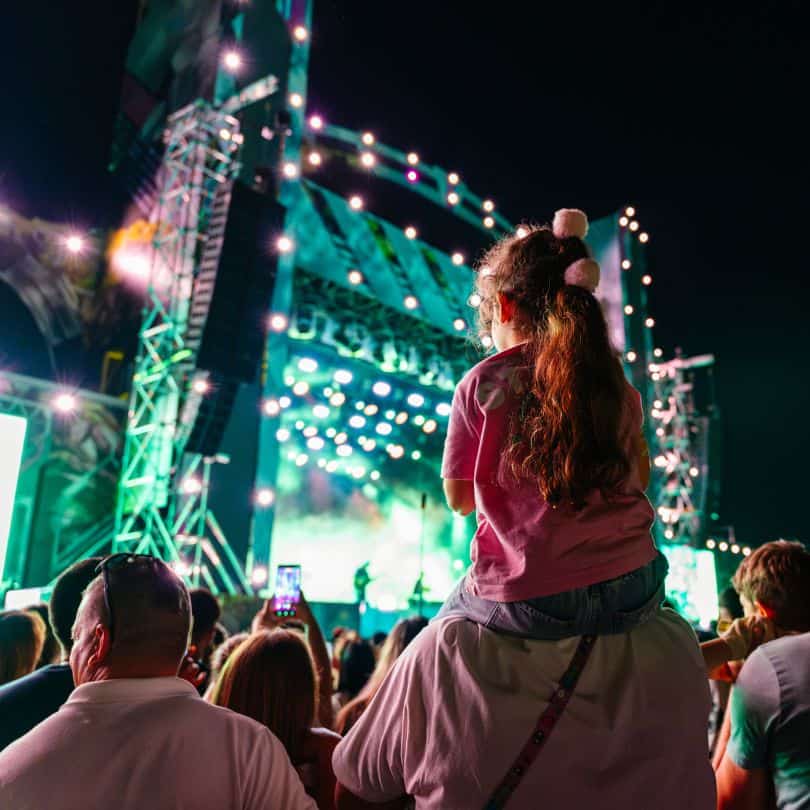 Fetiță pe umerii tatălui privind scena iluminată în timpul unui concert de noapte la Nibiru.